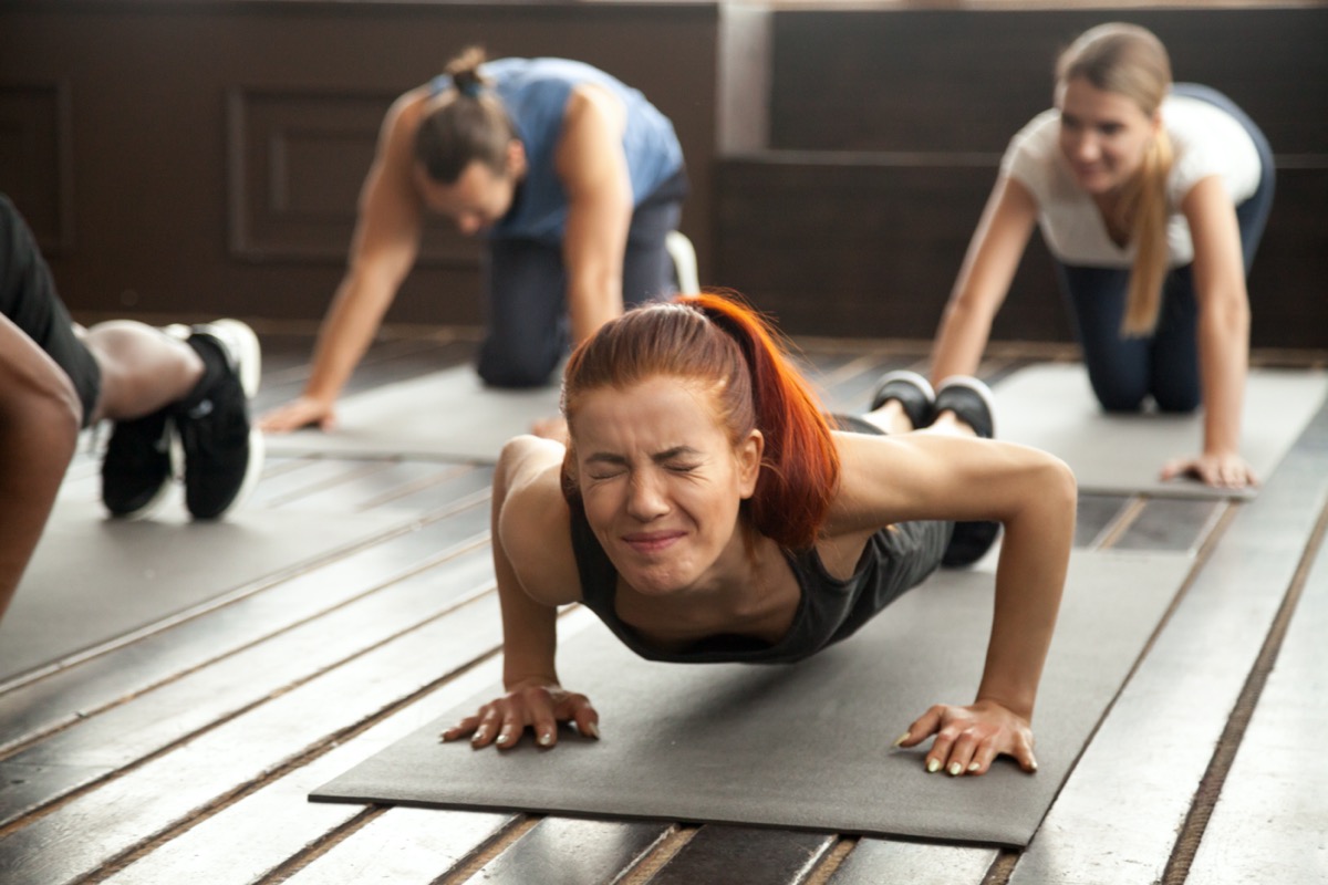 woman with painful expression on face doing difficult plank fitness exercise or push ups feeling sore muscles in diverse group training class at gym