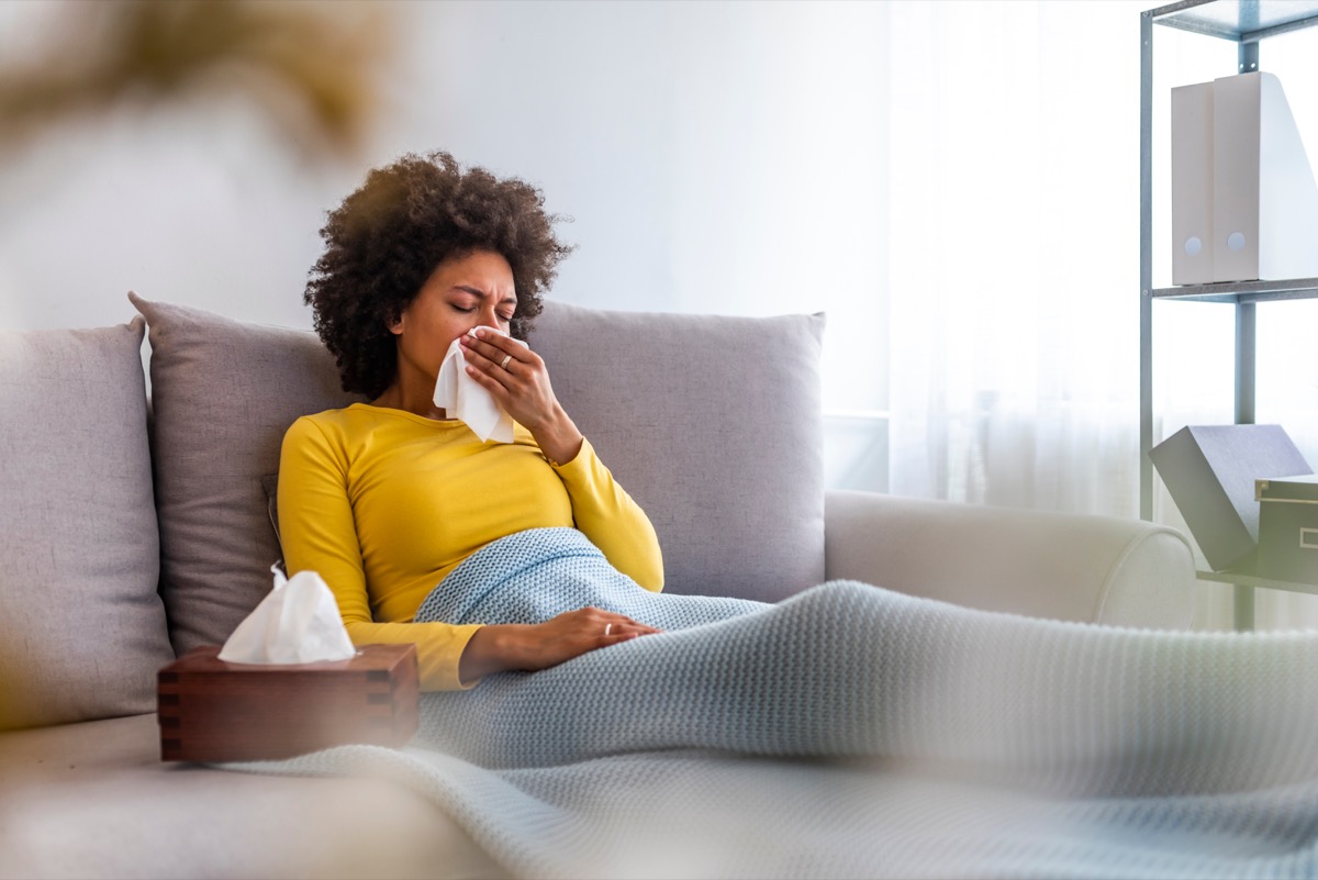 Woman sneezing into a tissue in the living room.  Woman blowing her nose on the couch at home in the living room.  African American woman wearing a headscarf sitting on a sofa at home