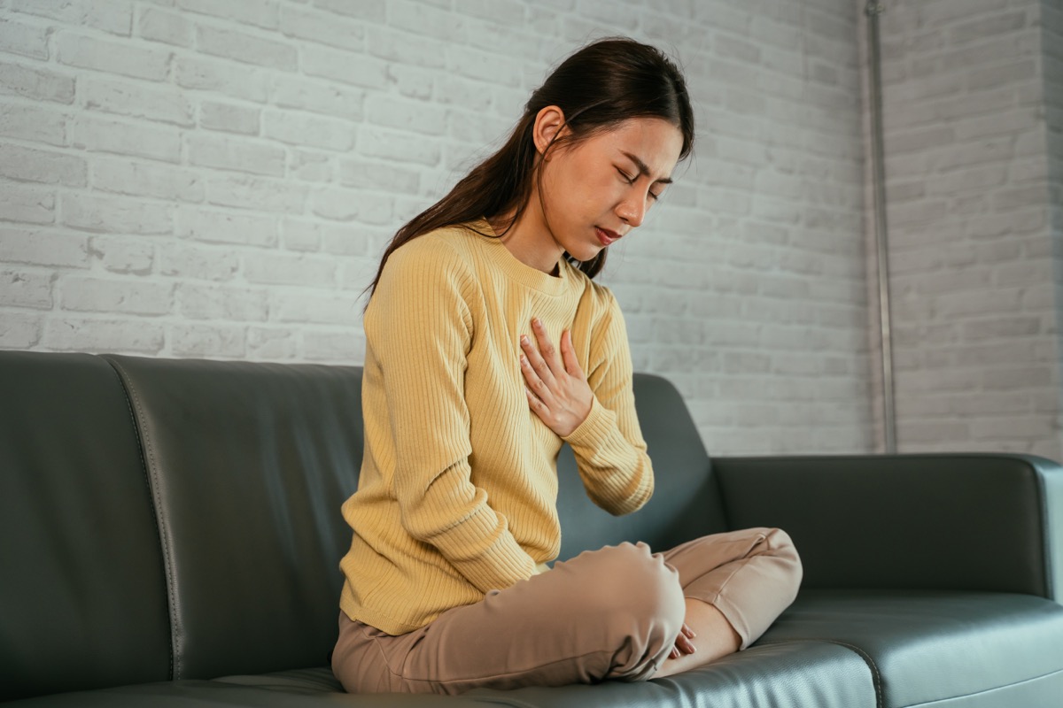 Asian young woman feeling uncomfortable because she is suffering from heartburn holding her chest with her eyes closed and sitting with her legs folded on the sofa at home.