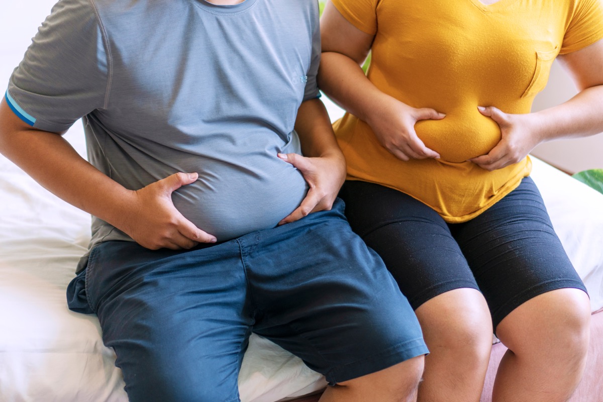 Overweight man and woman holding their bellies while sitting on bed.