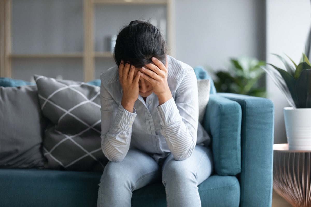 depressed indian woman holding head in hands, sitting alone on sofa at home