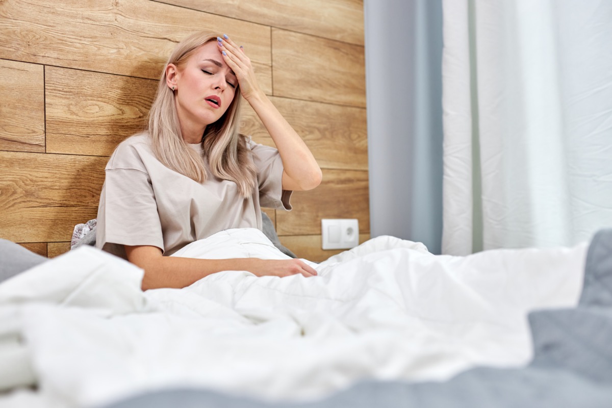 Sick woman with flu sitting in bed alone at home, having a high fever or temperature, touching her forehead