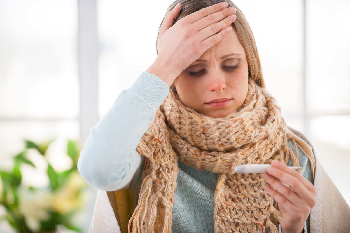 woman covered in plaid checking her body temperature while sitting on the bed in her apartment