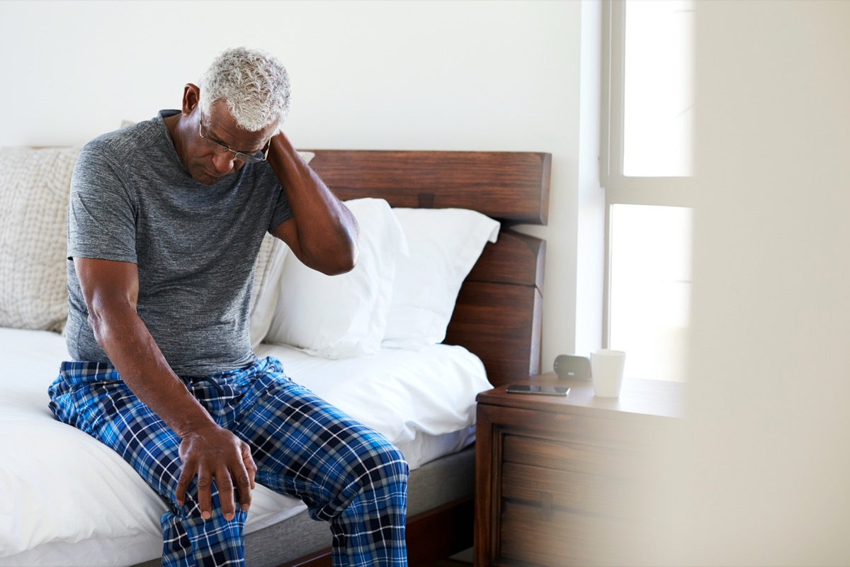 Senior man suffering from neck pain sitting next to bed at home