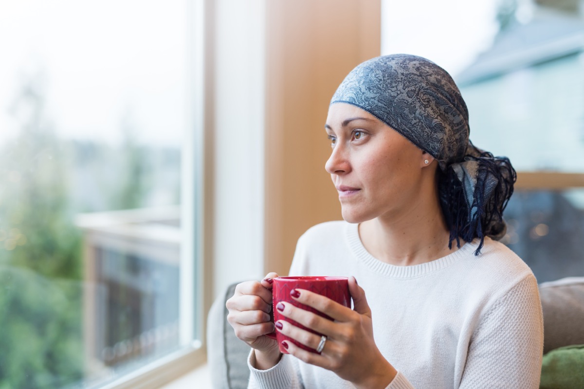 A woman in her 30s sits by her living room window with a cup of tea and looks out contemplatively.  She is a cancer survivor and wears a headscarf.