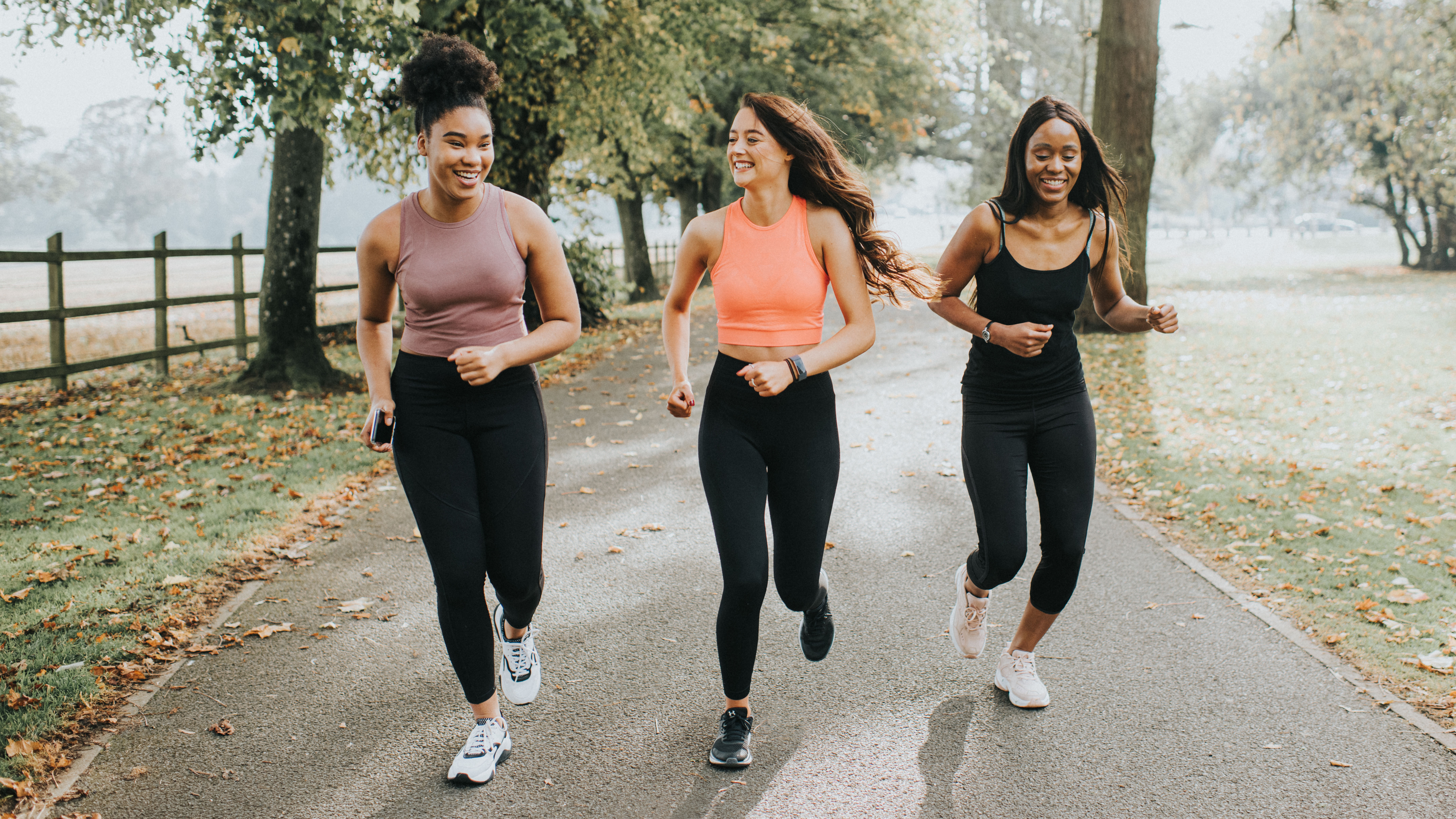 Three young women are running in a park;  there are leaves on the ground and fog in the background.