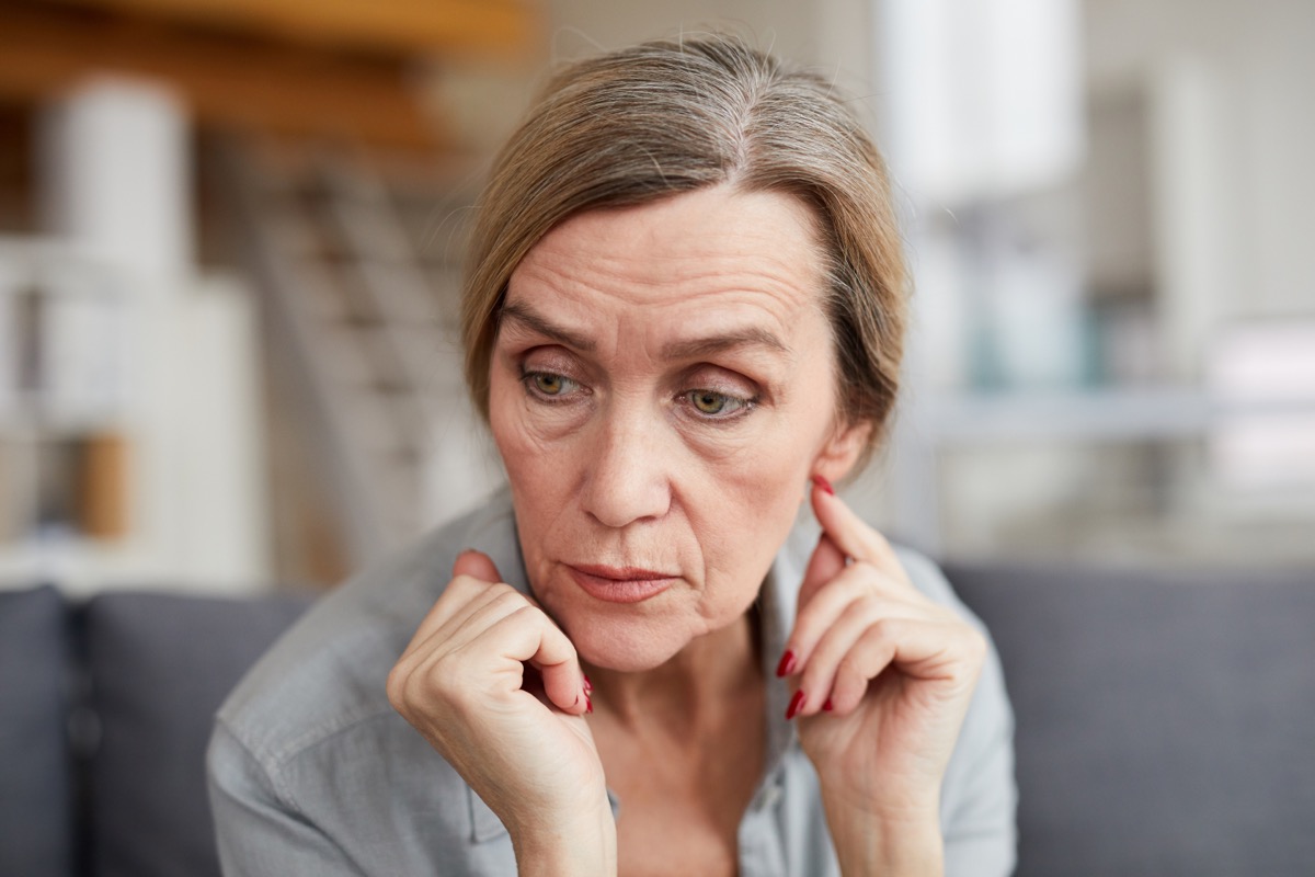 Portrait of sad mature woman sitting on sofa at home and looking away with concern and anxiety.