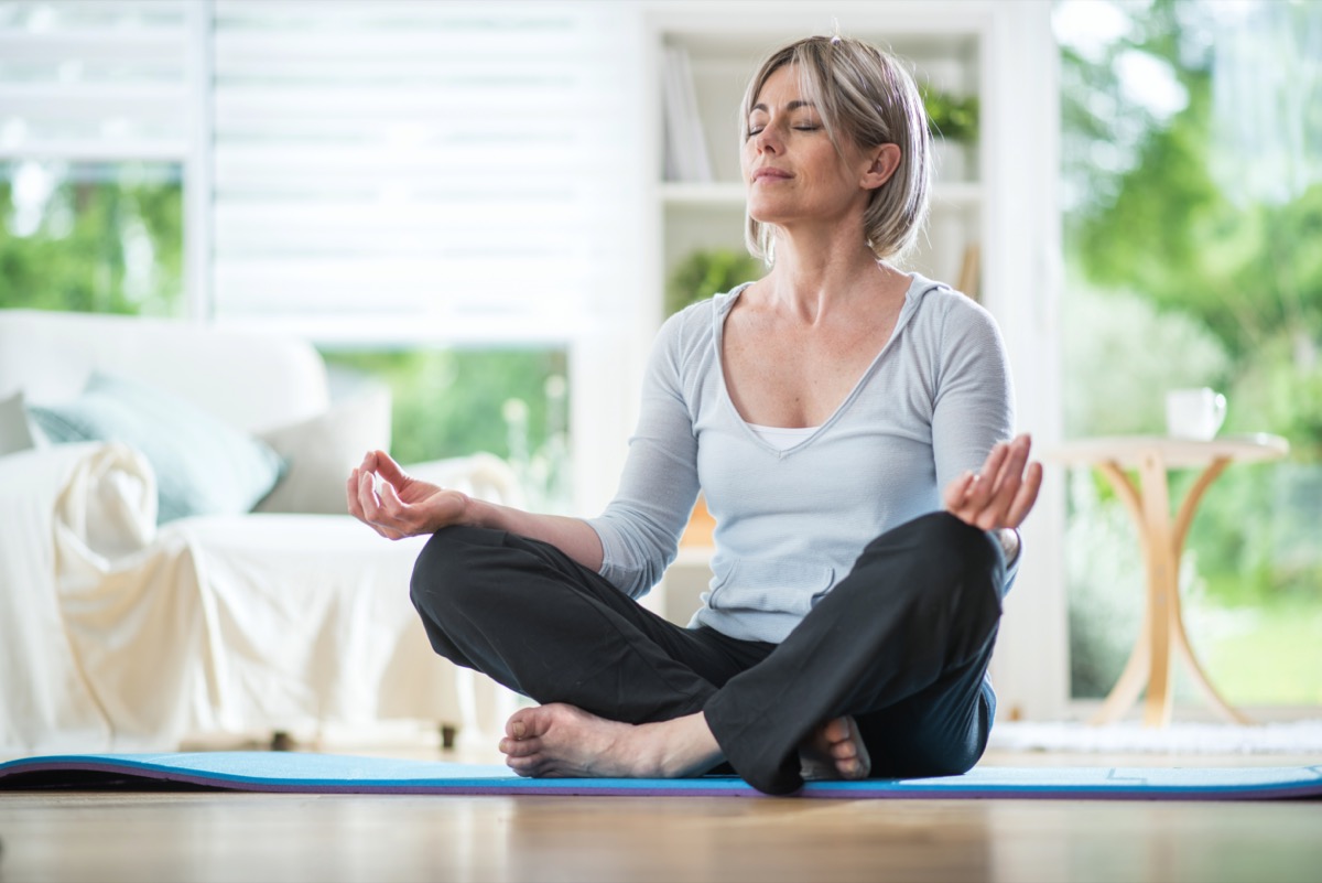 Middle-aged woman sitting in the lotus position on a rug in her living room.  his eyes are closed.  she is in the foreground