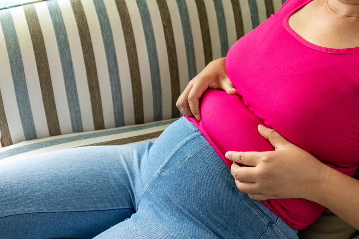 The woman shows excess fat around her waist while sitting on her sofa.