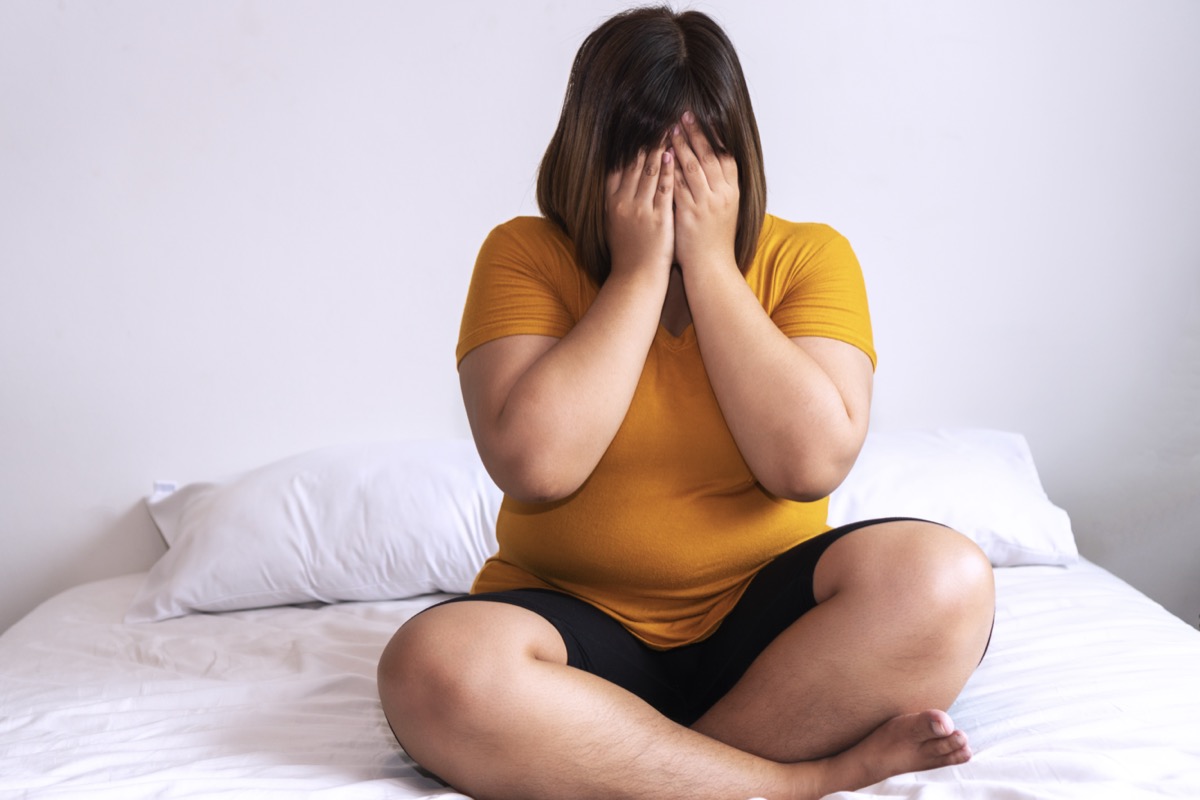 Overweight young woman sitting on a white bed while covering her face with her hands at home.  Upset woman suffering from extra weight in the bedroom.  Unhealthy obesity concept.