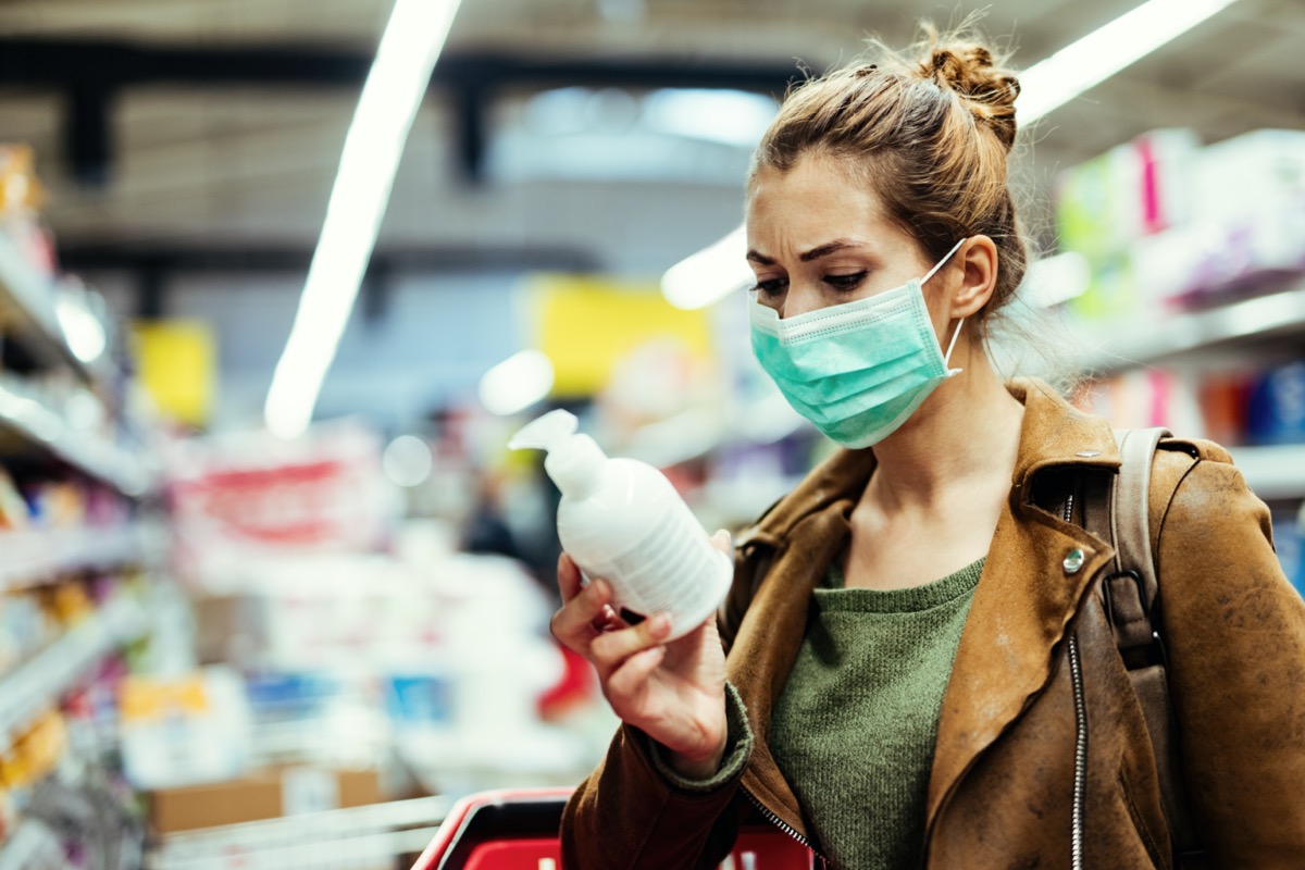 woman wearing protective mask reading label on bottle while buying hand soap in supermarket during virus epidemic