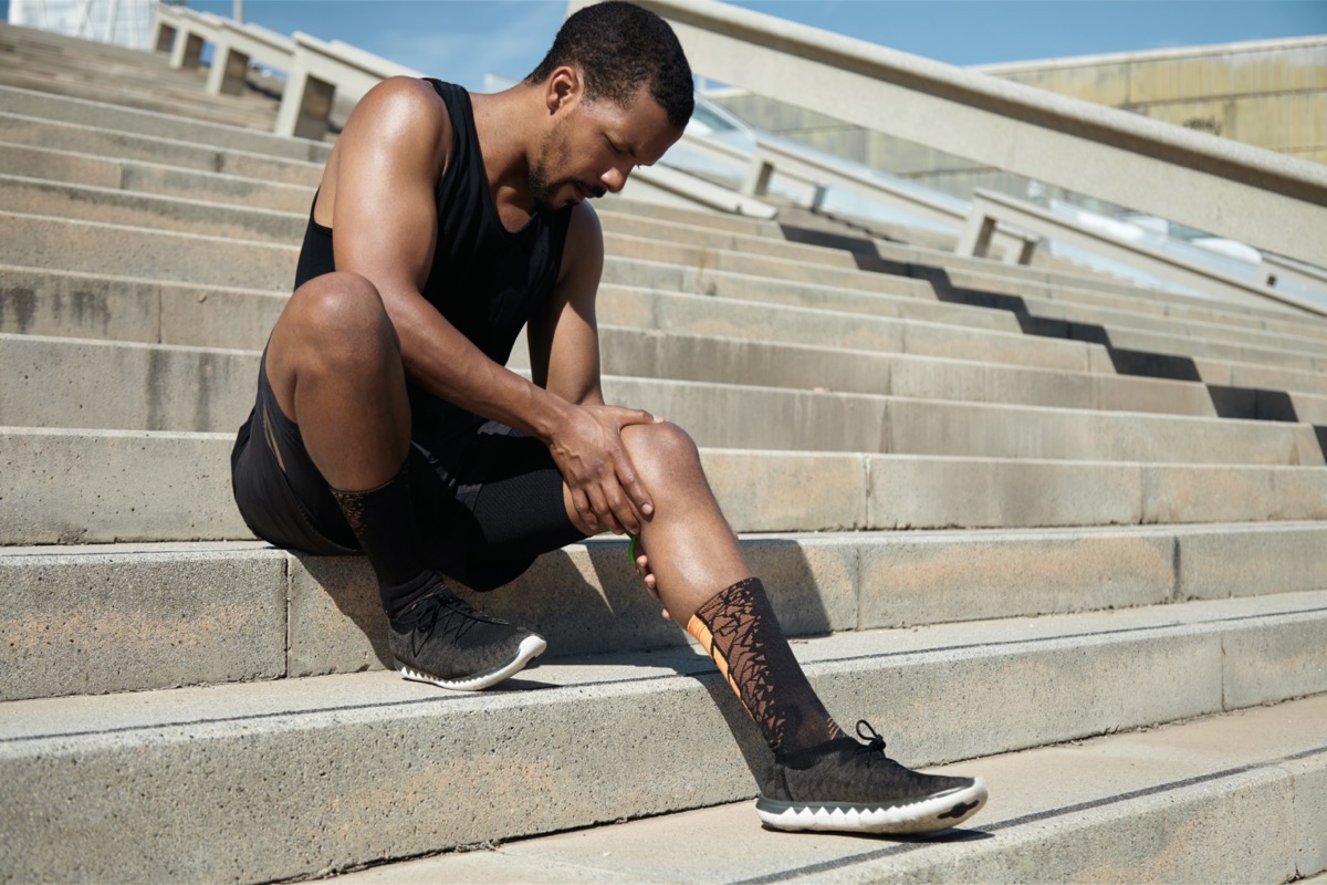 Black male runner in black sportswear and athletic shoes sitting on stairway outdoors clutching his sore knee