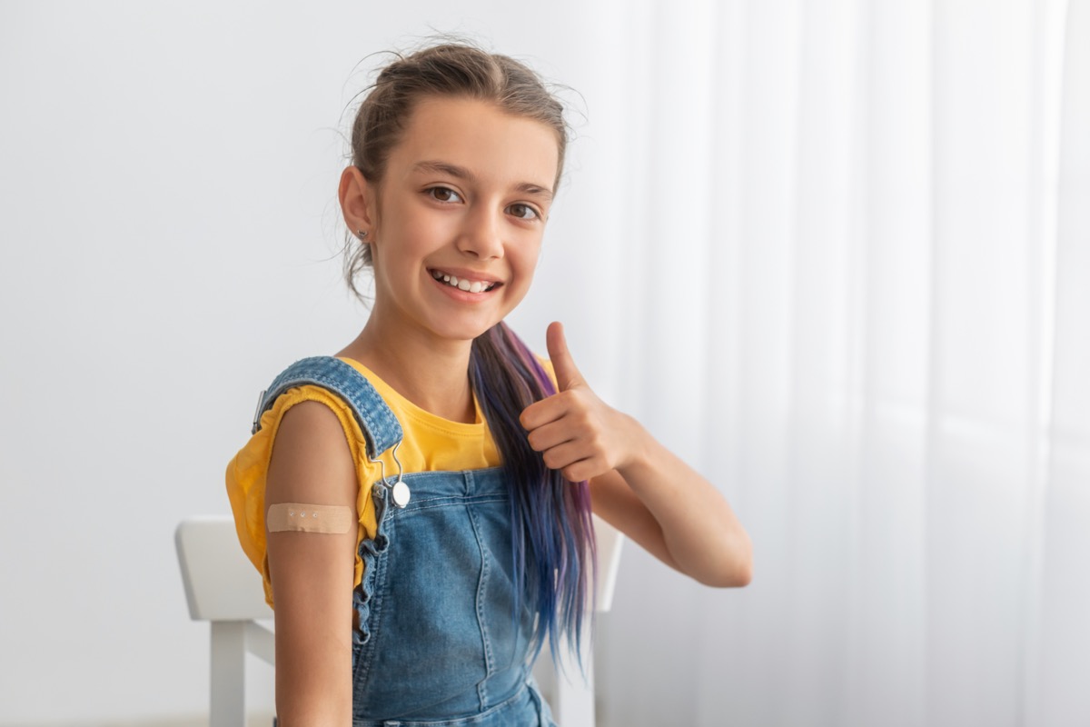 Cheerful smiling teenage patient showing vaccinated arm with patch sticking on shoulder after being shot and thumbs up gesture.