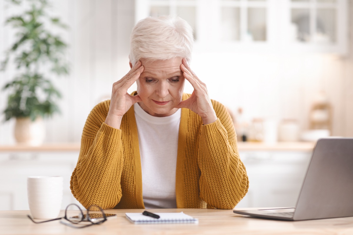 Senior lady taking notes, sitting in front of computer, touching her head