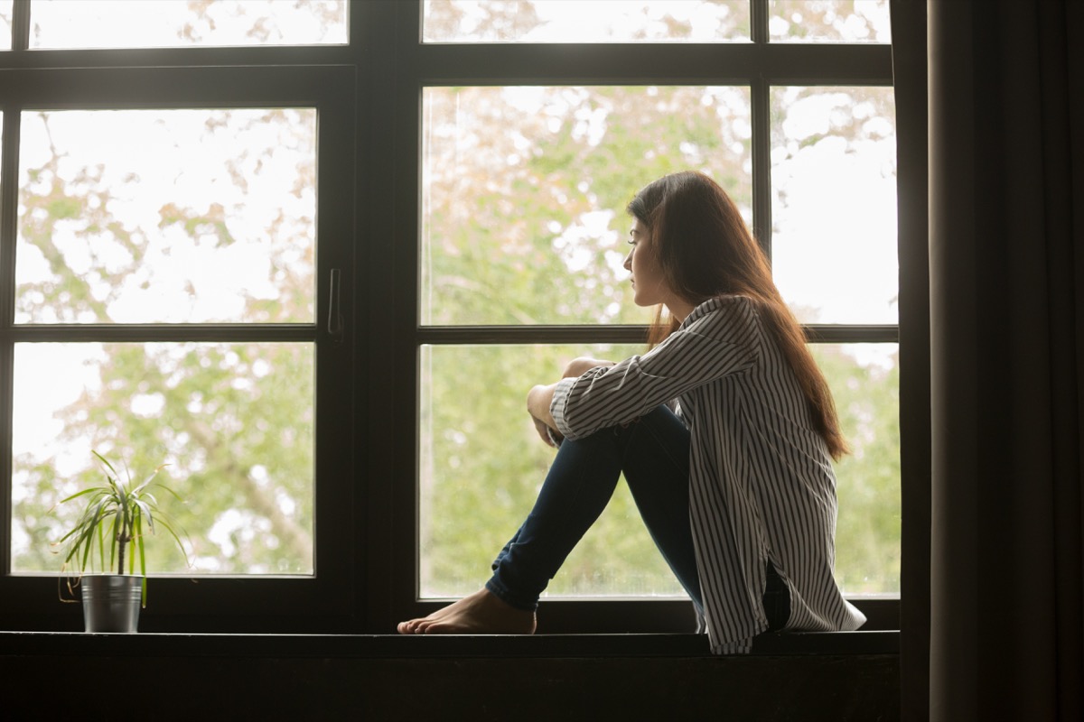 Thoughtful girl sitting on windowsill hugging knees looking at window, sad depressed teenager spending time alone at home, young upset pensive woman feeling lonely or frustrated thinking about problems