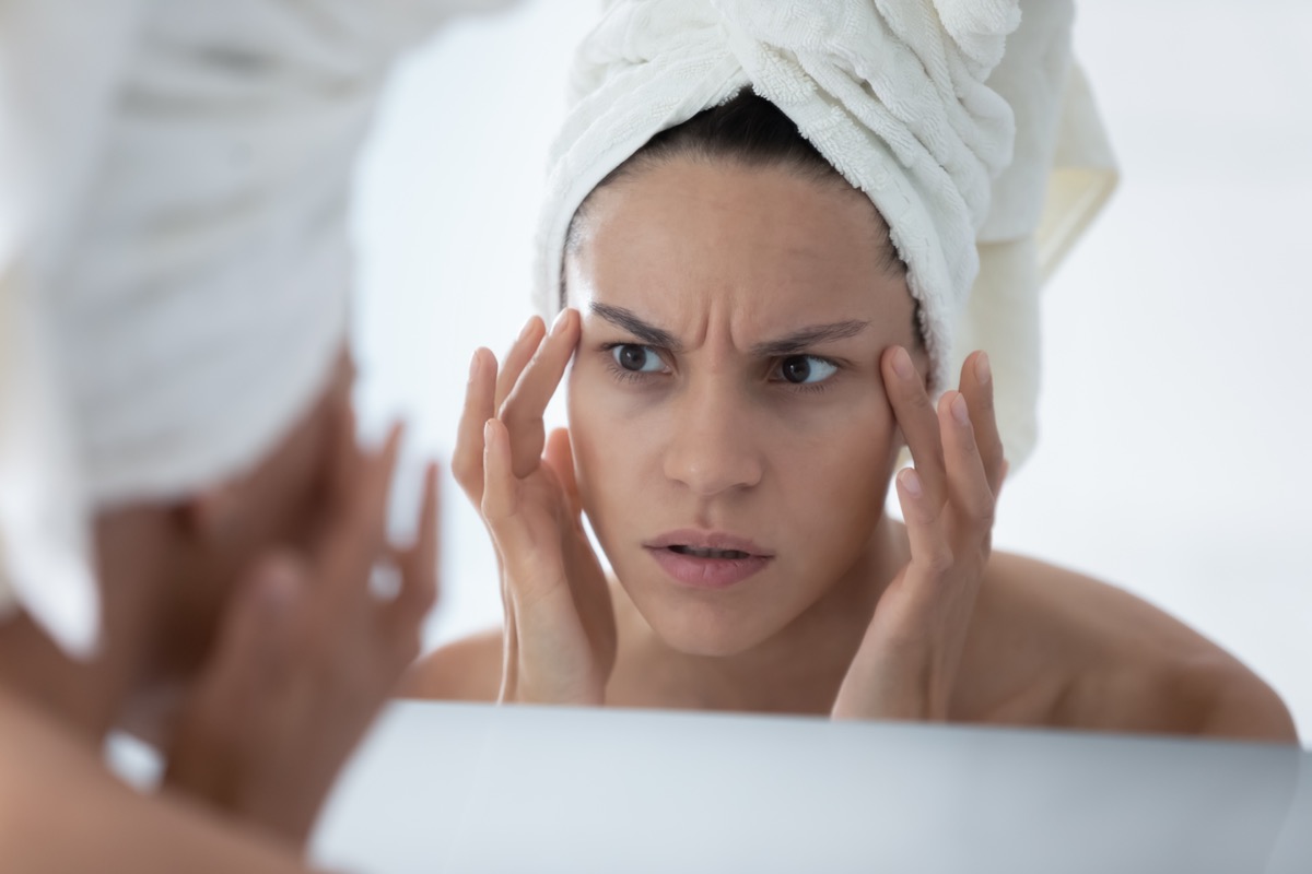 Unhappy woman in white bath towel checking skin after shower, looking in mirror, touching face skin.