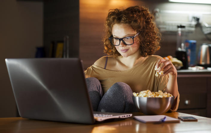 Young woman using computer and eating popcorn.  (Vesna Andjic/Getty Image)