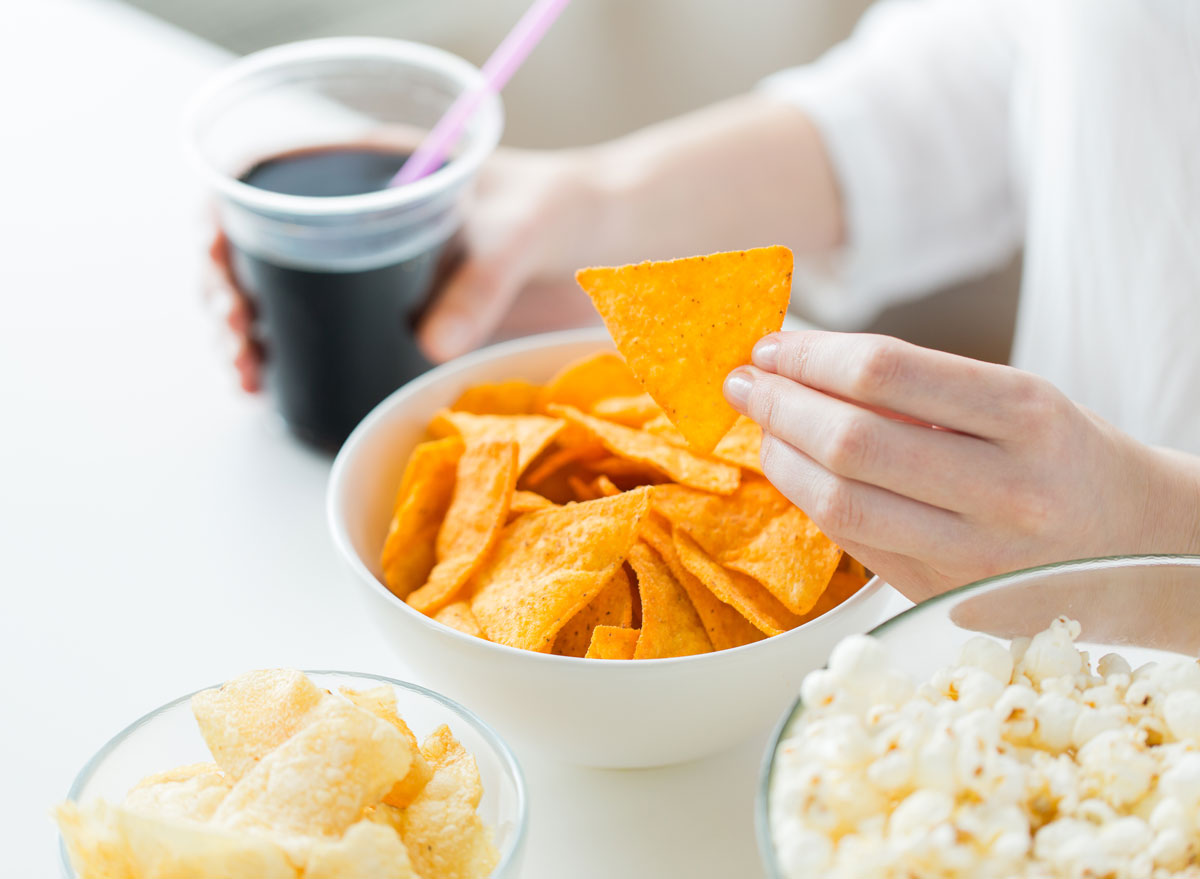 Woman looking for chips and holding soda in array of processed junk food at table with popcorn