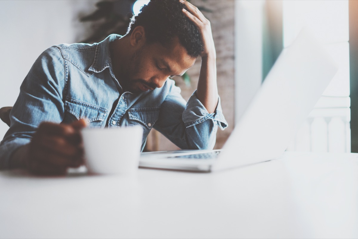 Tired young African man using laptop while sitting at the table on a sunny morning.  Concept of people working hard at home