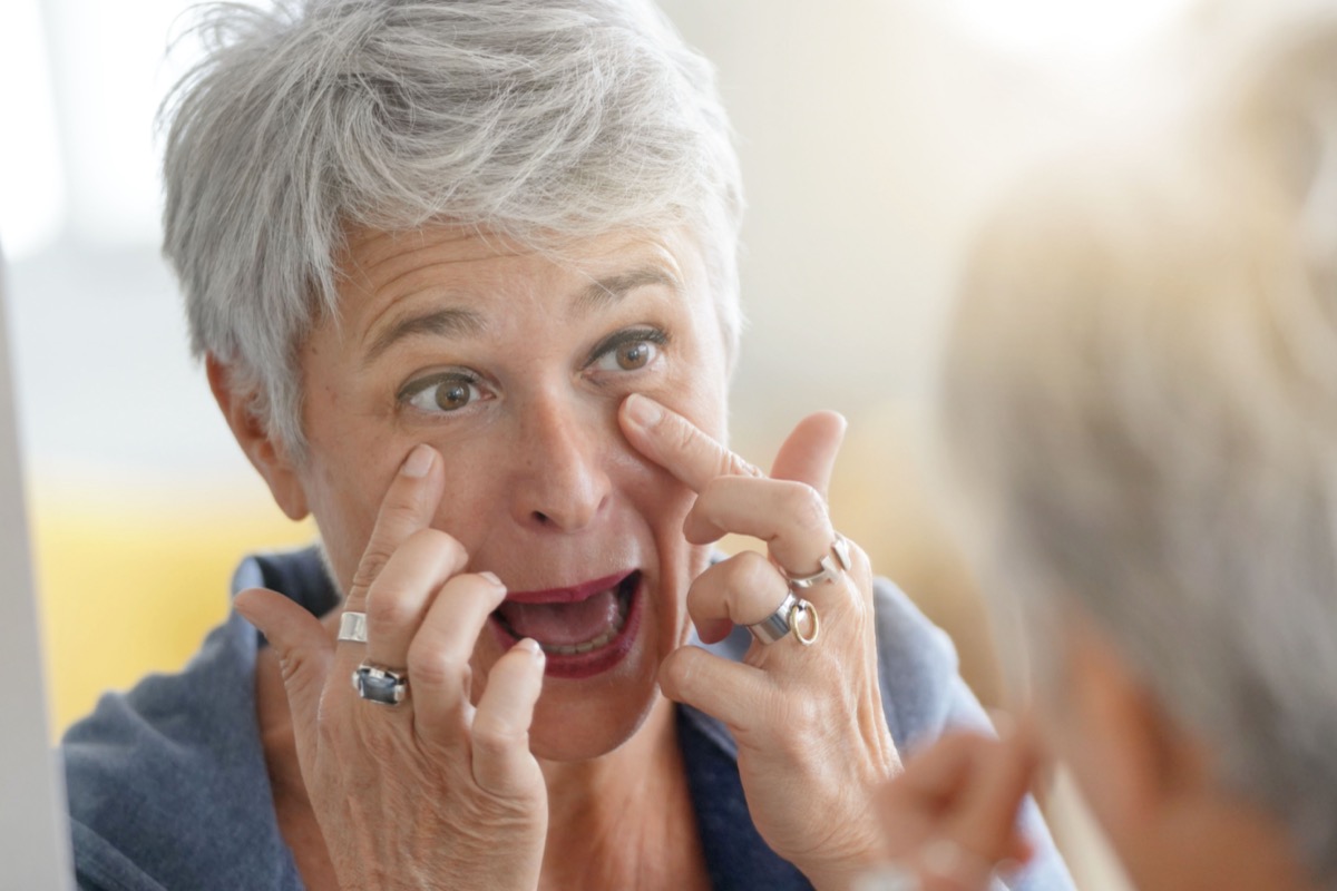 White-haired mature woman checking eye wrinkles in front of mirror.