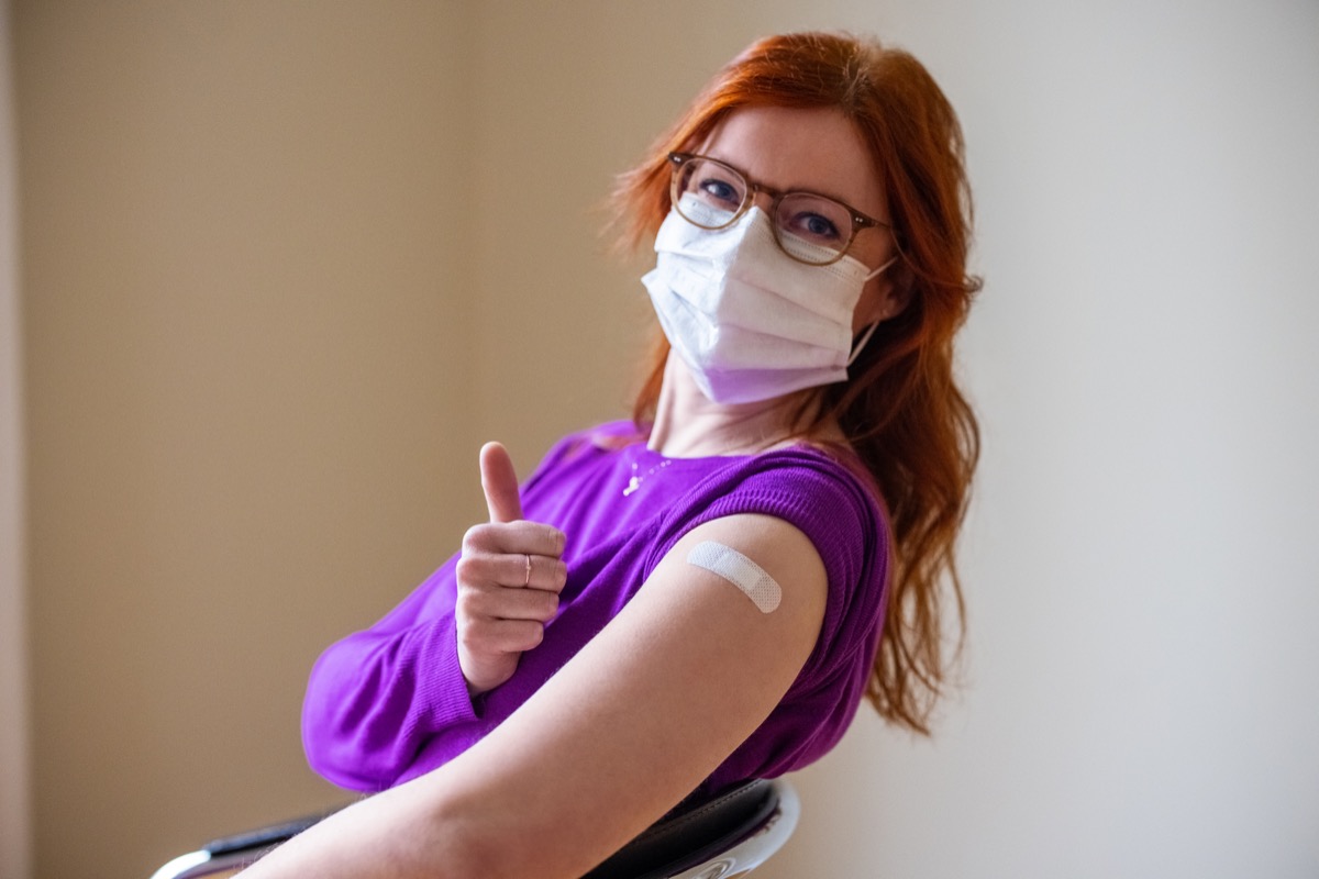 Woman wearing face mask looking at camera showing thumbs up after receiving covid-19 vaccine.