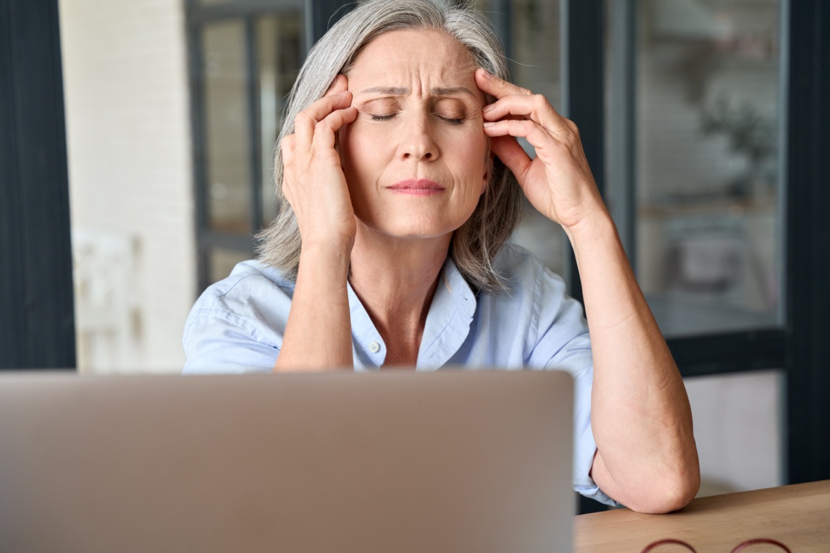 Stressed 60 years old working woman massaging head suffering from headache at home office.