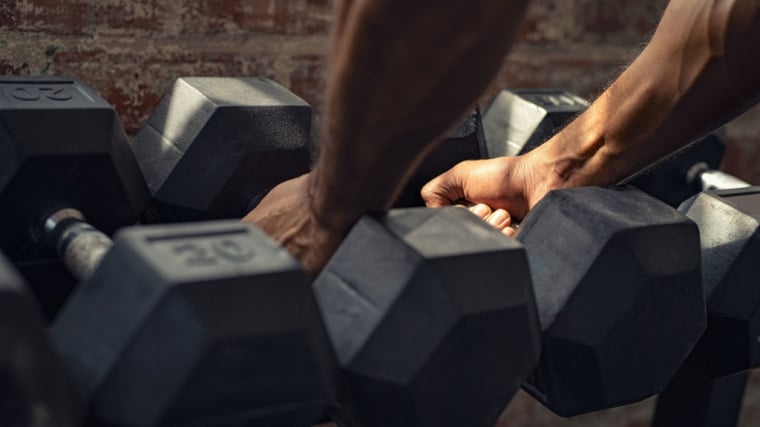 A close-up image shows a person's hands picking up dumbbells from a rack.