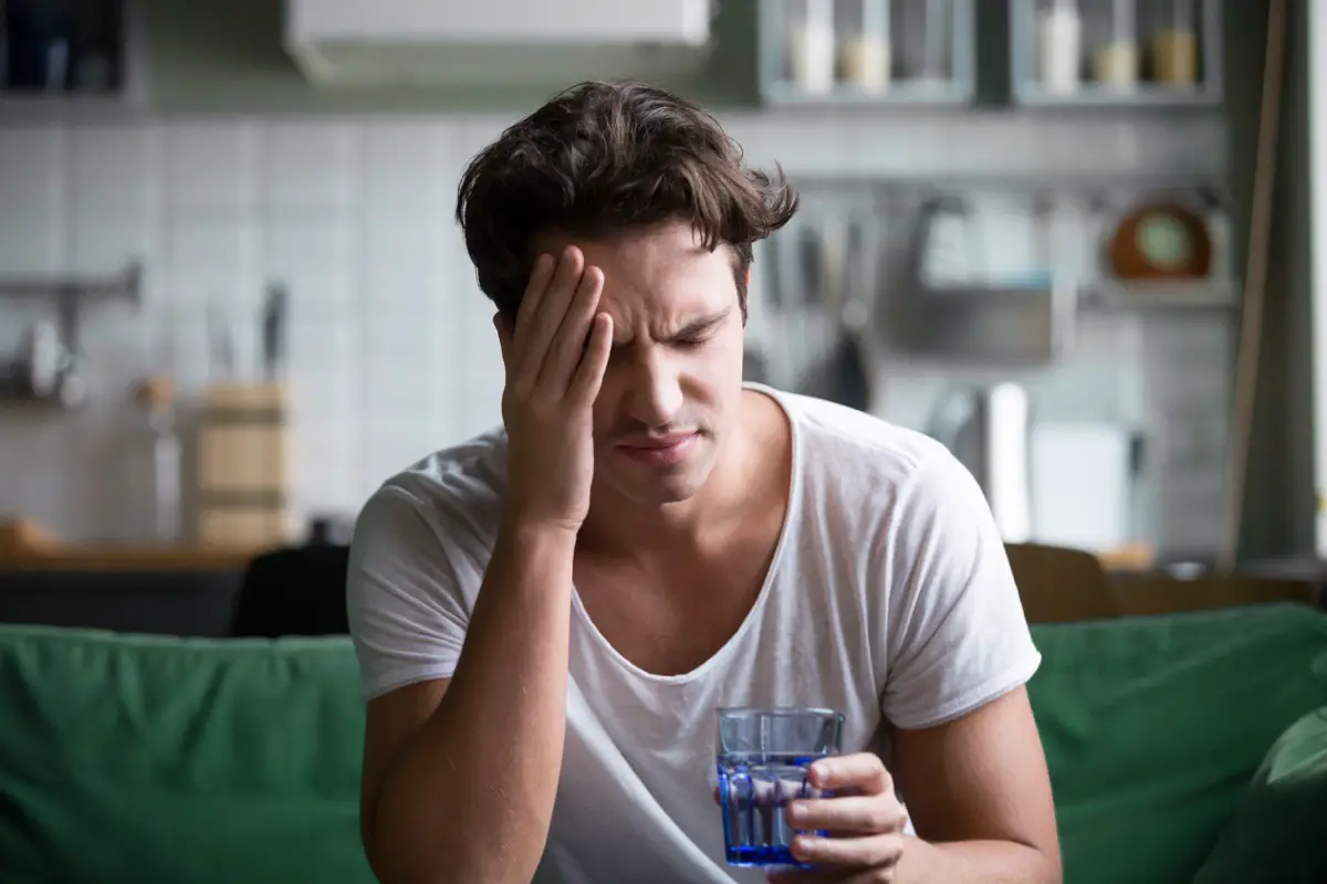 Young man suffering from strong headache or migraine sitting with glass of water in kitchen, millennial guy feeling intoxication and pain from touching sore head
