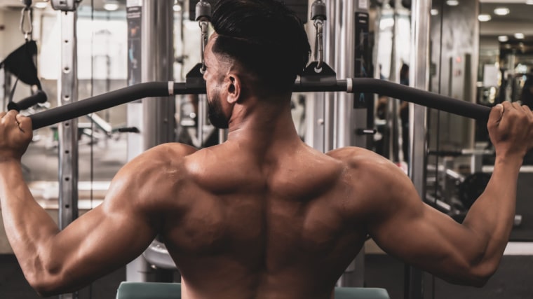 A shirtless person is featured from behind while they complete a lat pulldown in the gym.