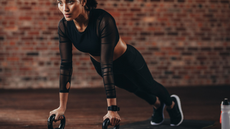 A person performs a plank on two kettlebells in a brick-walled gym.