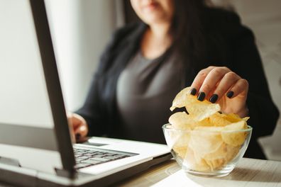 Binge eating, mindless snacking, junk food, unhealthy meals.  Woman eating potato chips from a bowl at her workplace